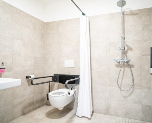 Barrierefreies Bad Komfort Gästehaus Dreibrunnen Barrierefreies Bad des Gästehauses Dreibrunnen. Eine Dusche, eine Toilette und Haltegriffe an der Toilette sind abgebildet. Das Bad ist beige-grau und hell ausgeleuchtet.