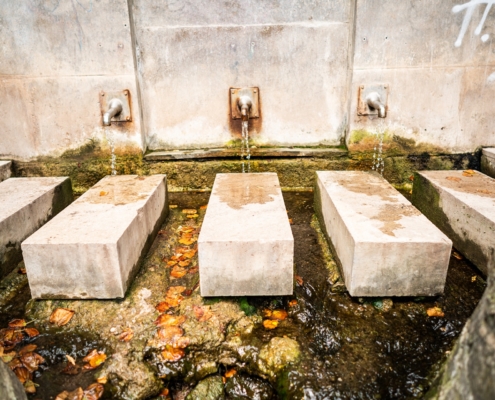 Aus einer Wand aus Stein entsprint die Dreibrunnen Quelle. Aus drei Wasserhähnen fließt jeweils eine Quellen in einen Fluss hinein.