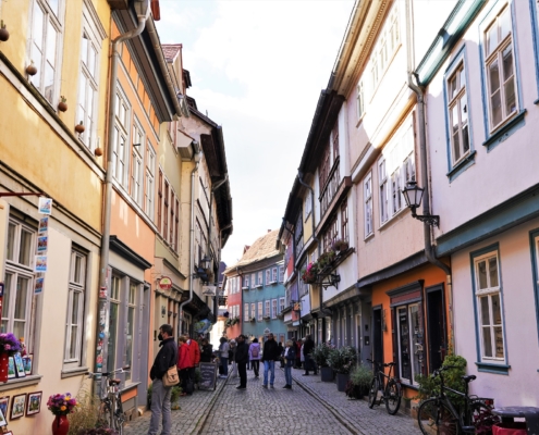 Die Krämerbrücke in Erfurt ist gesäumt von historischen Gebäuden, die sich auf der Brücke befinden. Zahlreiche Menschen genießen das Altstadtflair und schauen sich kleine Läden und Geschäfte an. Auf der Brücke stehen Blumenküble, die Gebäude sind ganz bunt.