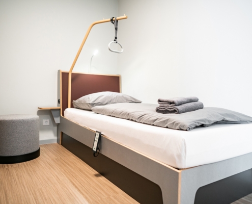 Komfort Zweibettzimmer barrierefrei Gästehaus Dreibrunnen Ein Pflegebett mit einem Haltegrifft über dem Bett. Neben dem Bett steht ein grauer Sessel. Auf dem Bett liegen zwei Handtücher die zusammengefaltet sind.
